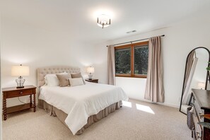 A bedroom with a large bed, two bedside tables with lamps, a window with curtains, and a mirror on the wall.