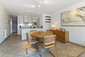 A modern kitchen with white cabinets, a central island, and a dining area with a wooden table and wicker chairs. - Plaza 309/0204 + P23* (SInt Idesbald)