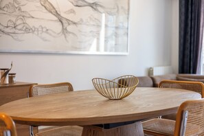 A round wooden dining table with a gold wire bowl centerpiece, surrounded by wicker chairs. - Plaza 309/0204 + P23* (SInt Idesbald)