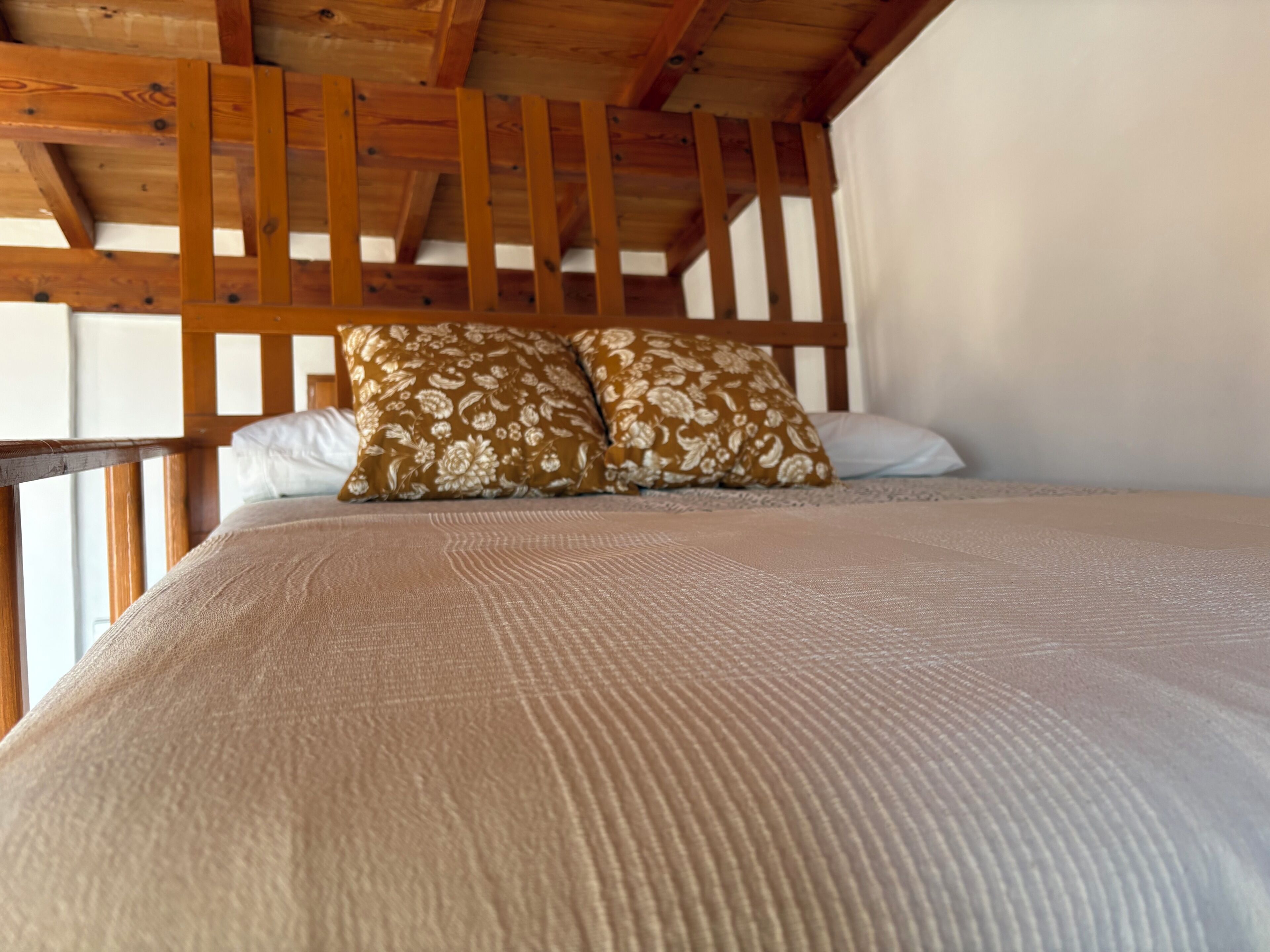 Un lit avec un oreiller à motifs et une couverture texturée, des poutres en bois au-dessus et un mur blanc.