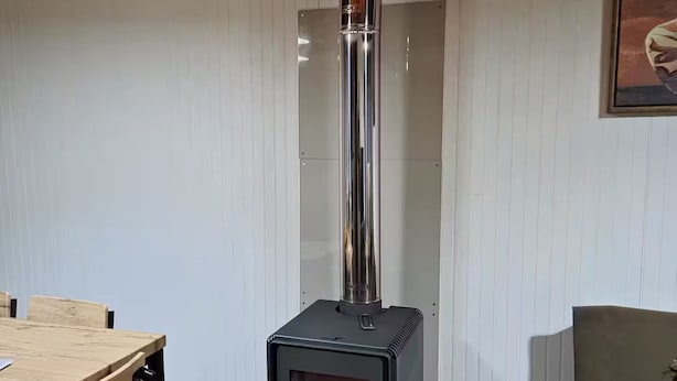 Een kamer met een zwart houthakkerkachel, een houten eettafel met stoelen, en een bank.