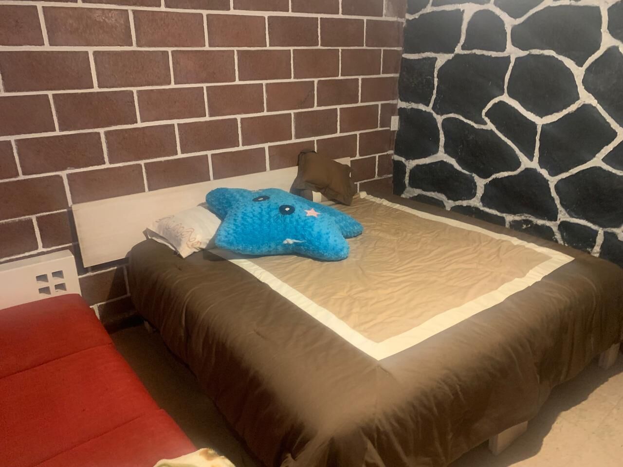 A bedroom with a brick-patterned headboard, a bed with a brown bedspread, a blue plush toy, and a red chair.