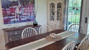 Een eetkamer met een donker houten tafel, witte stoelen, een kroonluchter, een kast en een schilderij van een kustlandschap.