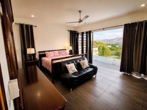 A bedroom with a bed, a black leather sofa, a wooden nightstand, a lamp, and a view of the outdoors. - Casa Alegre 5-bedroom 5 Bath Luxury home stunning ocean view awaits your retreat (San Juan Del Sur)