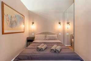 A neatly made bed with rolled towels, a painting on the wall, and wall-mounted lights. - CRE12 [Loreto - Cathedral] • Design • Boho • Chic (Milano)