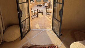 Un ventanal abierto que da a una terraza con una mesa y sillas, un tapete estampado y vistas del paisaje circundante.