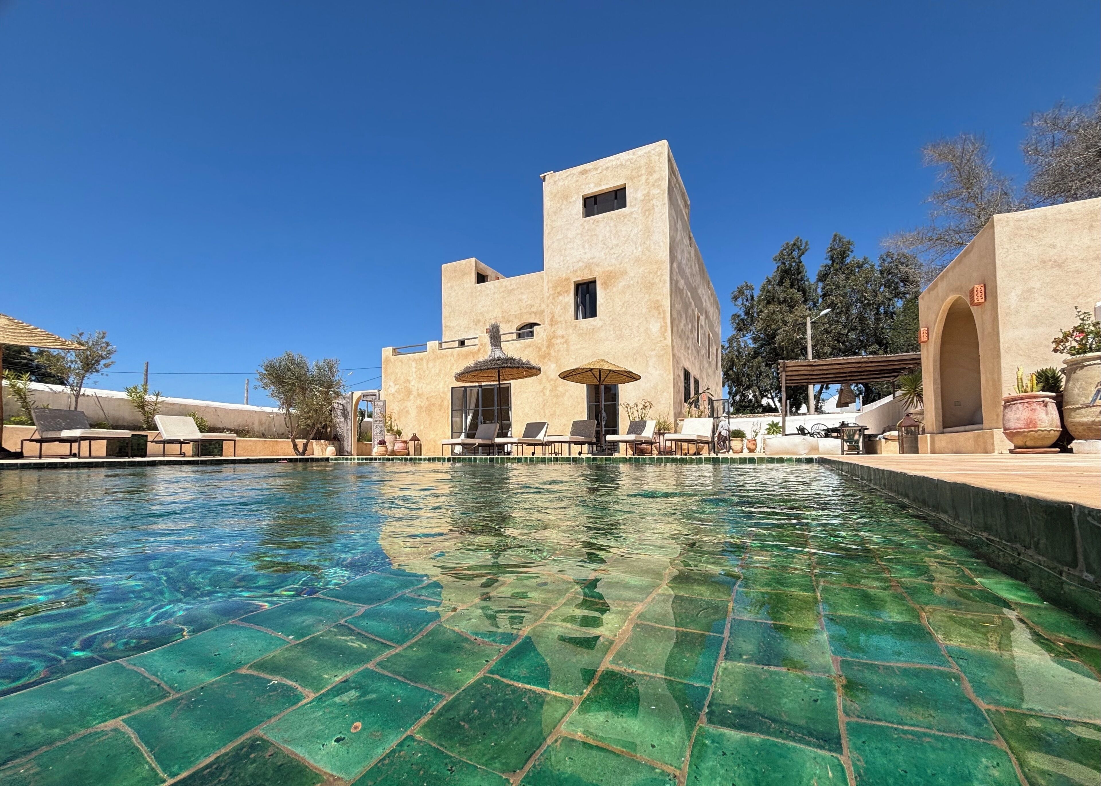 A pool area with lounge chairs and umbrellas, a building with a flat roof, and a clear blue sky.