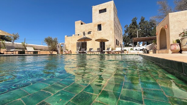 Zona de piscina con sillones y sombrillas, un edificio de techo plano y cielo azul despejado.