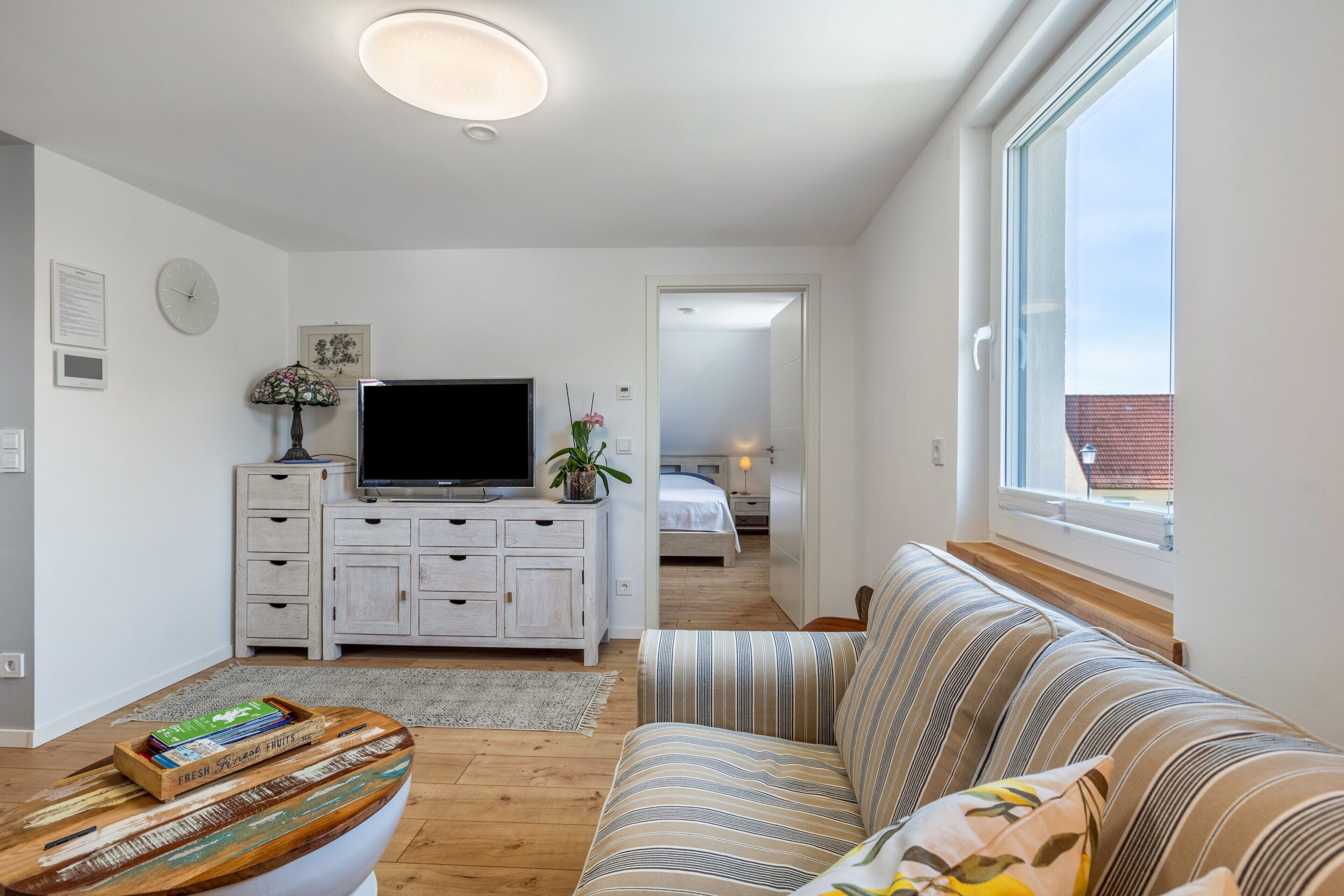 Ein modernes Wohnzimmer mit einer Couch, einem Fernseher auf einer weißen Kommode, einem Holztisch und einem Fenster mit Blick.