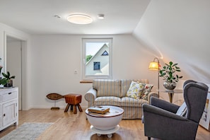 A modern living room with a striped sofa, a grey armchair, a round coffee table with a book, and a window with a view of a house.