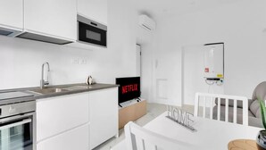 A modern kitchen with white cabinetry, a built-in oven, and a microwave. There is a dining area with a white table and chairs, and a television displaying 'NETFLIX' on the wall. - CRE9 [Loreto - Cathedral] • Central • Modern • (Milano)