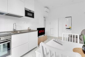 A modern kitchen with white cabinetry, a built-in oven, and a microwave. There is a dining area with a white table and chairs, and a television displaying 'NETFLIX' on the wall. - CRE9 [Loreto - Cathedral] • Central • Modern • (Milano)