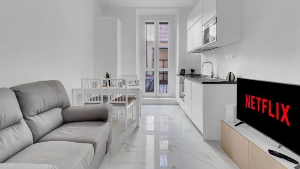 A modern living room with a sofa, a dining table, and a kitchen area with a TV displaying 'NETFLIX'. - CRE9 [Loreto - Cathedral] • Central • Modern • (Milano)