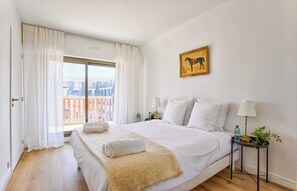 A hotel room with a large bed, a window with curtains, a small table with a lamp, and a painting on the wall. - Charming Apt w/ Large Balcony - 2bd/4p -deauville (Deauville)
