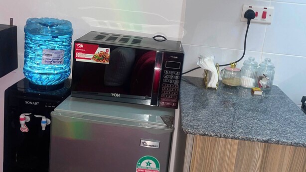 A kitchen with a water dispenser, a microwave, a refrigerator, and a wooden cabinet. - G.BlissHomes | Stylish Modern Studio Near Nairobi CBD with King Bed & Work space (Nairobi)