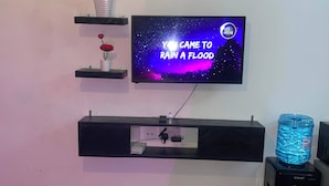 A living room with a mounted TV displaying a space-themed show, a water dispenser, and a plant on a shelf. - G.BlissHomes | Stylish Modern Studio Near Nairobi CBD with King Bed & Work space (Nairobi)