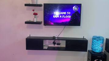 Un salon avec un téléviseur fixé au mur diffusant une émission sur le thème de l’espace, un distributeur d’eau et une plante posée sur une étagère.