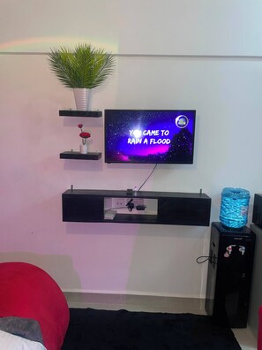 Ein Wohnzimmer mit einem an der Wand montierten Fernseher, der eine Weltraumshow zeigt, einem Wasserspender und einer Pflanze auf einem Regal.