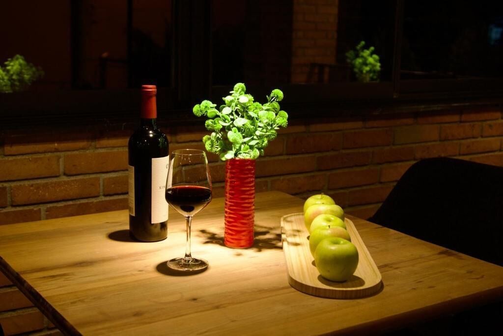 A table with a bottle of wine, a glass, a red vase, and a tray with green apples.