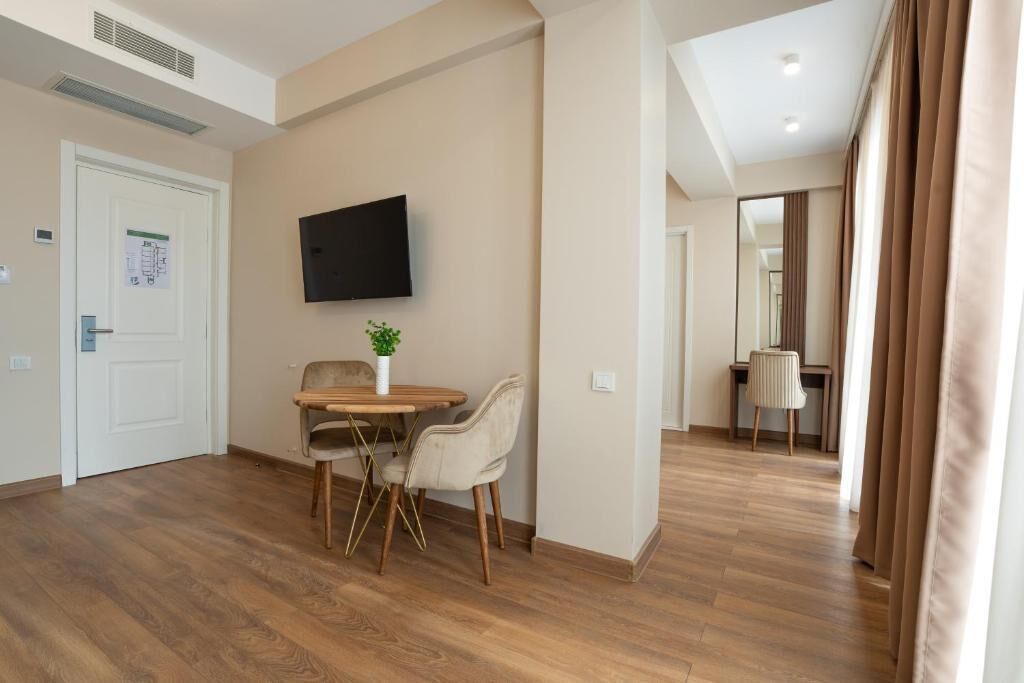 A hotel room with a wooden floor, a dining table with chairs, a flat-screen TV mounted on the wall, and a door with a sign.