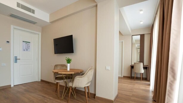 Una camera d'hotel con pavimento in legno, un tavolo da pranzo con sedie, un televisore a schermo piatto montato a parete e una porta con un cartello.