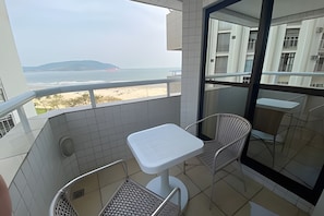 Uma varanda com mesa e cadeira brancas, vista para a praia e o mar, e chão de azulejos.