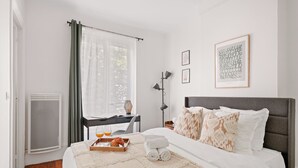 Ein Hotelzimmer mit Bett, Schreibtisch mit Stuhl, Fenster mit Vorhängen, Heizkörper und einem gerahmten Bild an der Wand.