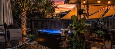 A hot tub surrounded by outdoor furniture, a wooden fence, and a sunset sky.
