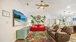 Ein Wohnzimmer mit einem roten Sofa, einem braunen Liegestuhl, einem farbenfrohen Teppich, einem Deckenventilator und einer Wand mit tropischen Kunstwerken.