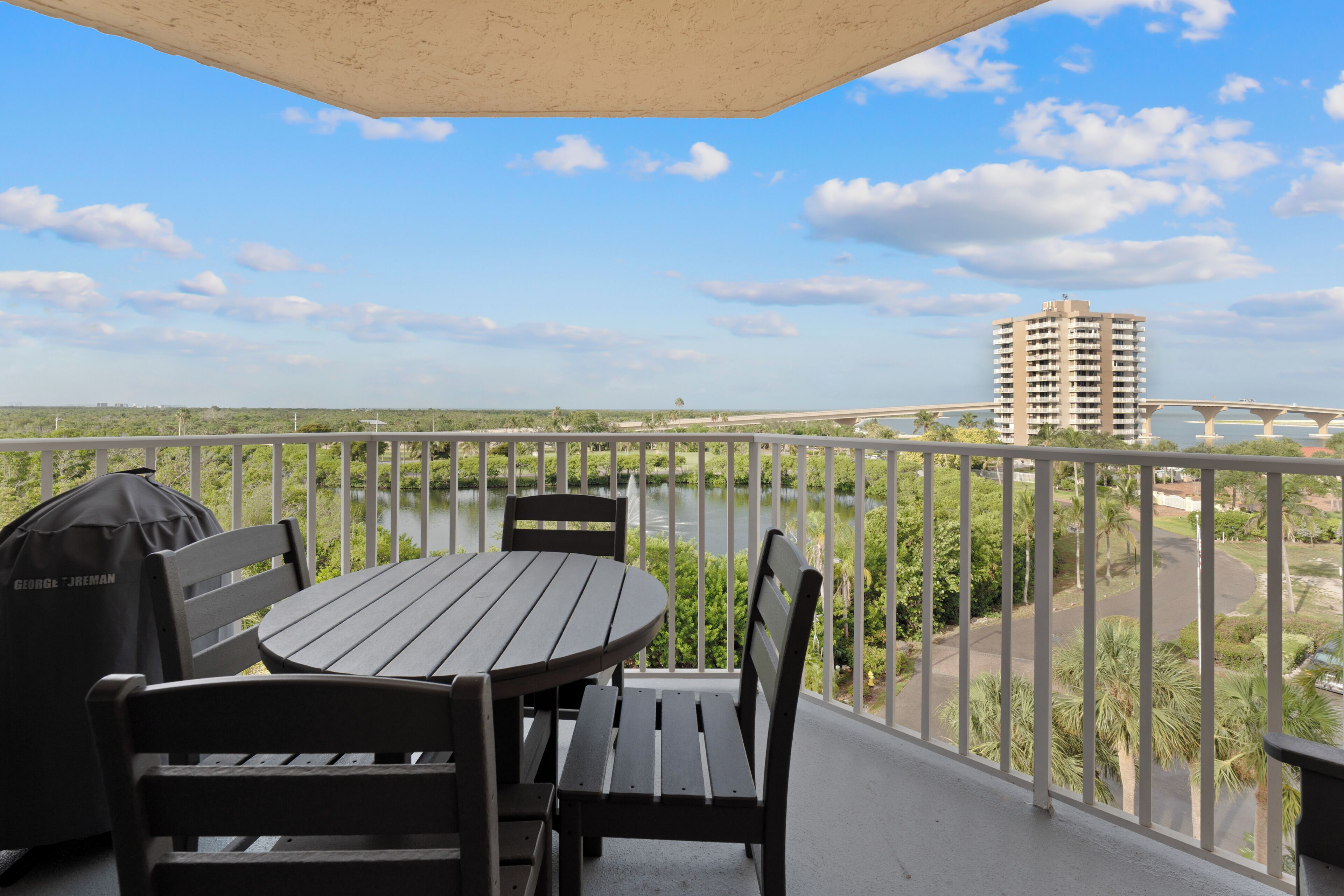 A balcony with a table and chairs, a grill, and a view of a bridge and greenery.