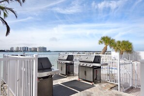 Eine Außenpatio mit zwei Grillstationen, einem weißen Zaun und Blick auf das Meer und Gebäude im Hintergrund.