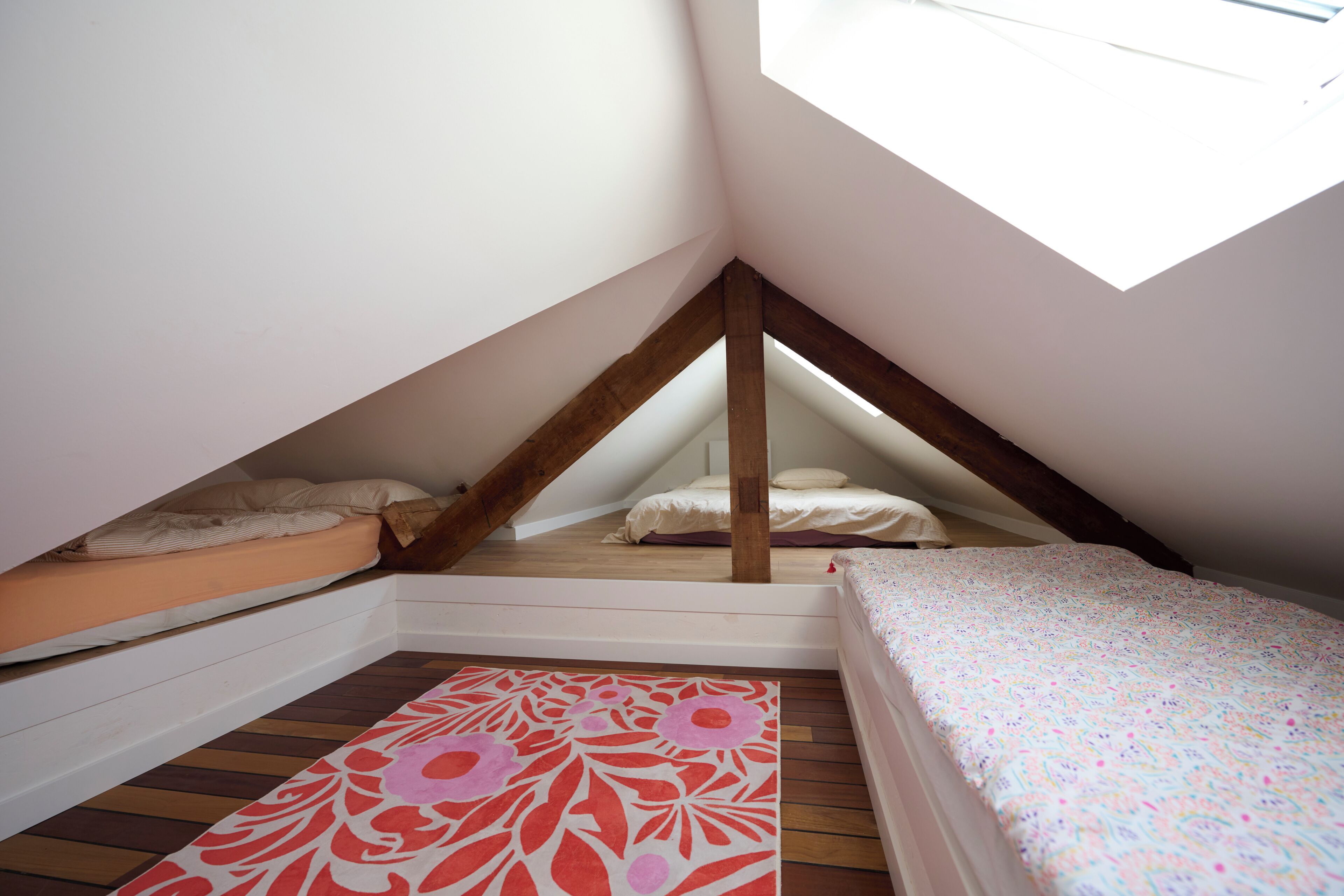 Une petite chambre mansardée avec deux lits, une lucarne, des poutres en bois et un tapis à motifs.