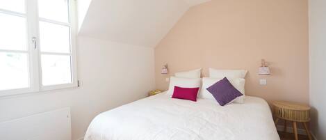 Quarto com teto inclinado, cama com roupa de cama branca, duas luminárias de cabeceira e uma pequena mesa de cabeceira de madeira.