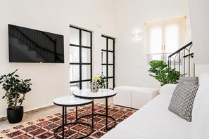 A modern living room with a white sofa, a round table, a TV mounted on the wall, and a staircase with a railing. - Kaletzia Villa IV - Four Bedroom Villa, Sleeps 8 (Zakinthos)
