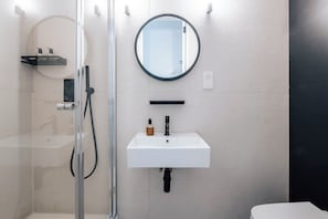 Ein Badezimmer mit einer gläsernen Duschkabine, einem weißen Waschbecken mit schwarzem Wasserhahn und einem runden Spiegel an der Wand.