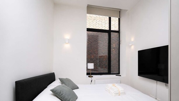 Een slaapkamer met een bed, kussens, een raam met uitzicht op een bakstenen gebouw, een tv aan de muur en een radiator.