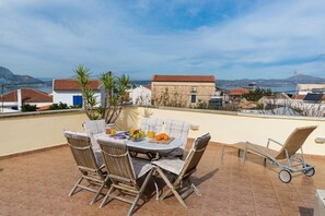 A patio with a table set for breakfast, surrounded by wicker chairs, overlooking a coastal landscape with buildings and mountains. - Villa Epavlis - Three Bedroom Villa, Sleeps 6 (Plaka)