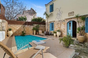 A pool area with lounge chairs, a stone wall, and a building with a sign. - Villa Epavlis - Three Bedroom Villa, Sleeps 6 (Plaka)