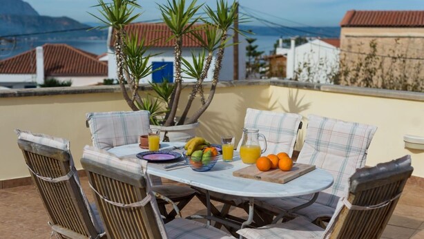 Una mesa al aire libre con sillas, un mantel, una jarrón, un vaso de jugo de naranja y un bol de fruta.