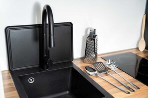 Eine Küche mit einem schwarzen Spülbecken, einem Wasserhahn, einer Reibe, einem Pfannenwender, einem Löffel und einer Zange.