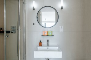A bathroom with a glass shower enclosure, a white sink, a mirror, and a shelf with toiletry items. - The Antigallican by Nestor | Cosy studio (London)