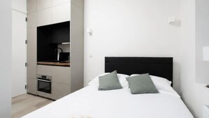 A bedroom with a bed, pillows, a nightstand, a built-in desk with a lamp, and a small kitchenette. - Cosy Victorian studio near The O2 & Greenwich (London)