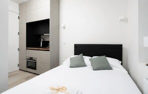 Ein Schlafzimmer mit einem Bett, Kissen, einem Nachttisch, einem integrierten Schreibtisch mit Lampe sowie einer kleinen Küchenzeile.