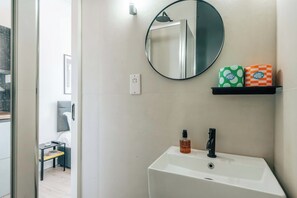 A bathroom with a white sink, a round mirror, a showerhead, and a shelf with toiletries. - The Antigallican by Nestor | Cosy studio (London)