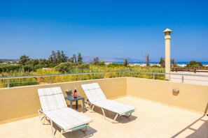 Ein Balkon mit zwei Liegestühlen, einem kleinen Tisch, einer Flasche und einem Glas, und Blick auf das Meer und die Berge.