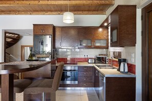 A kitchen with wooden cabinets, a built-in oven, a sink, and a dining area with a chair and table. - Villa Portokalia - Two Bedroom Villa, Sleeps 4 (Platanias)
