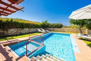 A rectangular swimming pool with a red safety railing, surrounded by a stone wall and a grassy area with lounge chairs. - Villa Portokalia - Two Bedroom Villa, Sleeps 4 (Platanias)