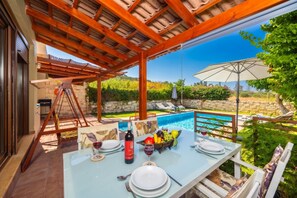 An outdoor dining area with a pool, wooden pergola, and stone wall backdrop. - Villa Portokalia - Two Bedroom Villa, Sleeps 4 (Platanias)