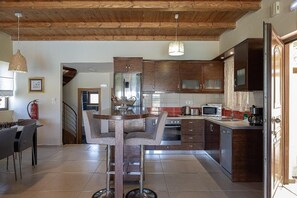 A modern kitchen with wooden cabinets, a central island, and a dining area with a chair. - Villa Lemonia - Two Bedroom Villa, Sleeps 4 (Platanias)