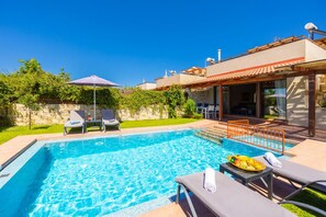 A swimming pool with lounge chairs and a table, surrounded by a stone wall and greenery. - Villa Mandarin - Two Bedroom Villa, Sleeps 4 (Platanias)